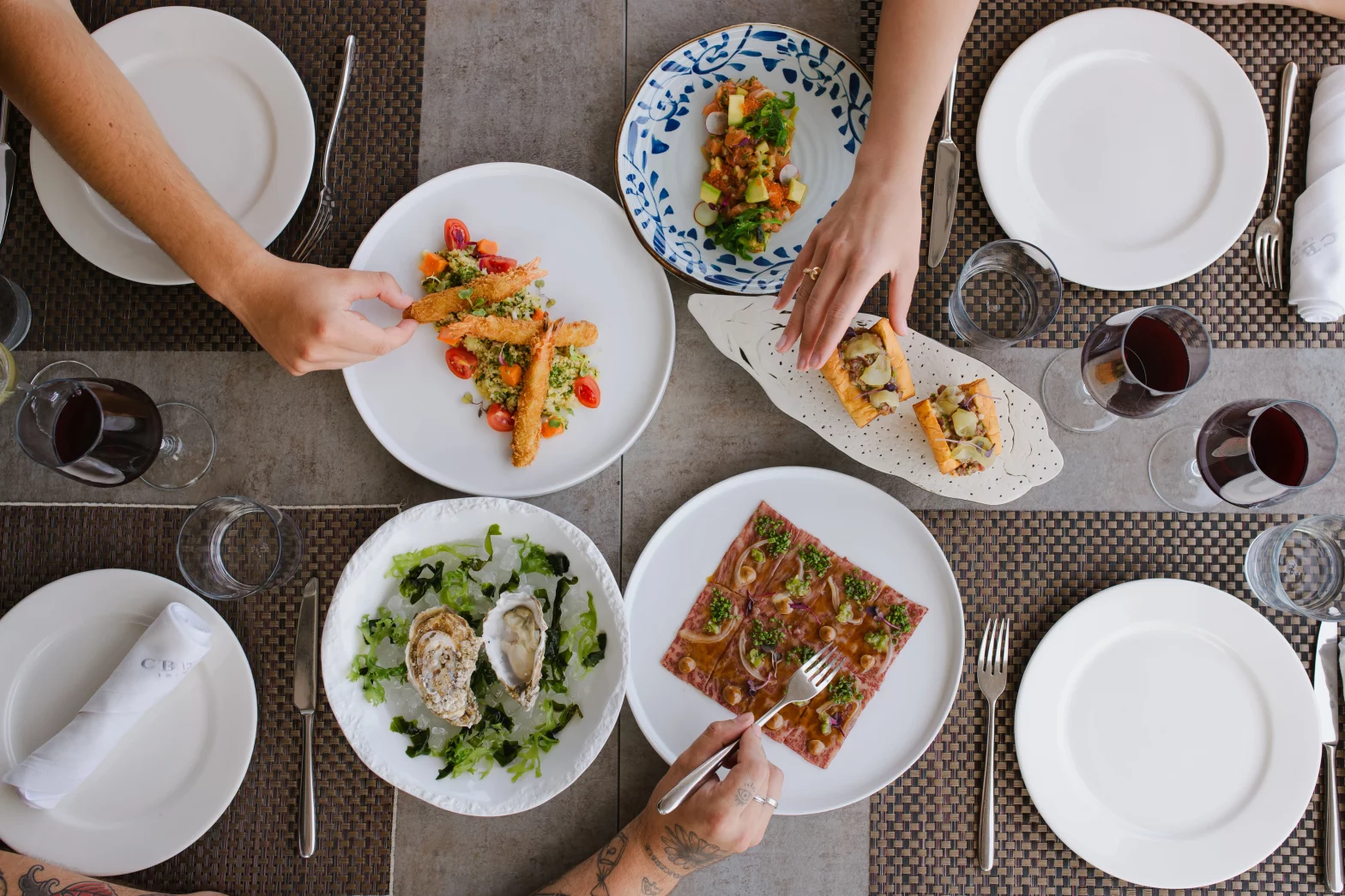 Una mesa servida con diferentes platos degustación con manos que cogen la comida. Restaurante CBbC Marina Sta. Eulalia